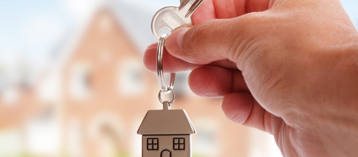 landlord's hand holding a house key in front of a rental property